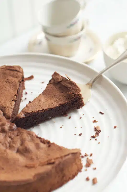 slice of queen of sheba cake (reine de saba) on white cake stand with tea cups in the background
