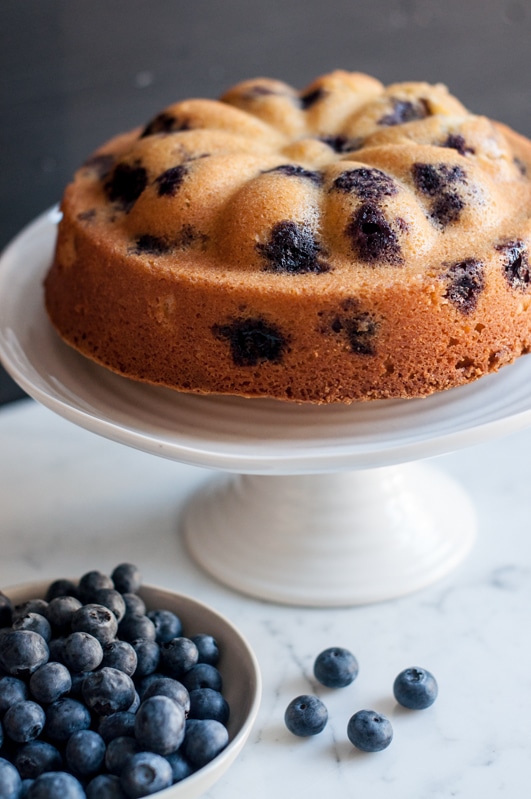 blueberry lemon cake on white cake stand with fresh blueberries in bowl
