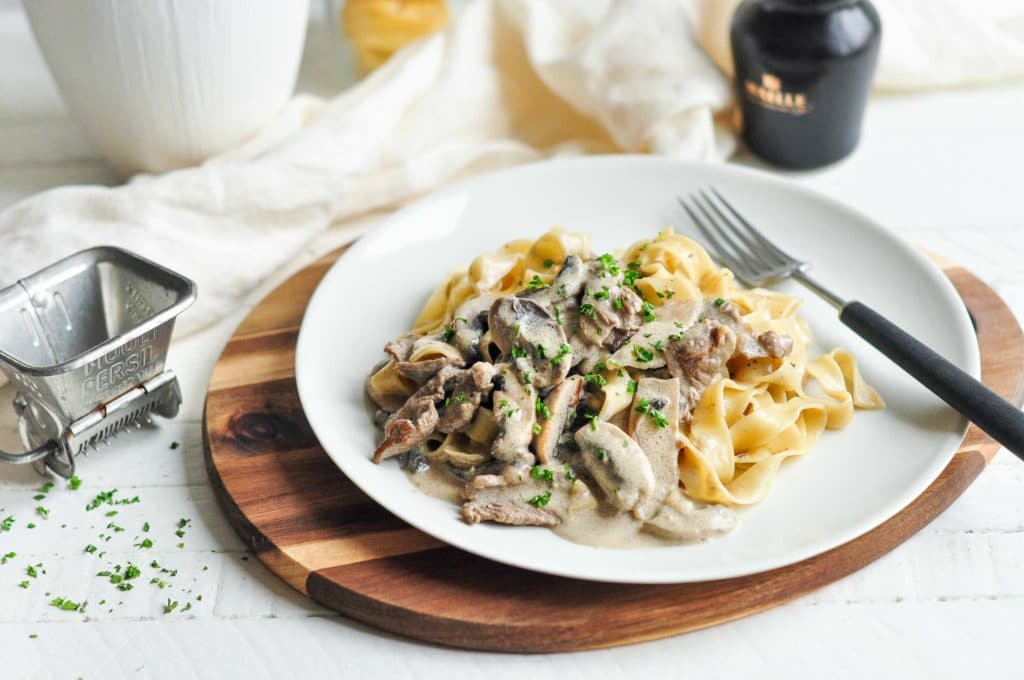 beef stroganoff with pasta on beige plate with fork and herb grinder