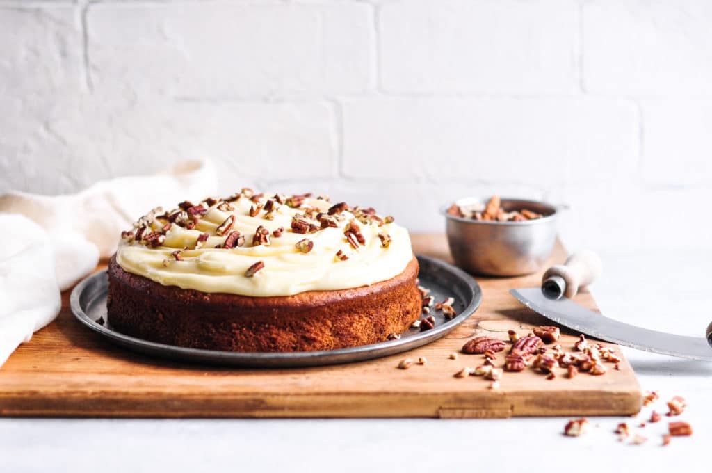 easy banana cake decorated with cream cheese frosting and chopped pecans on metal tray and wooden board