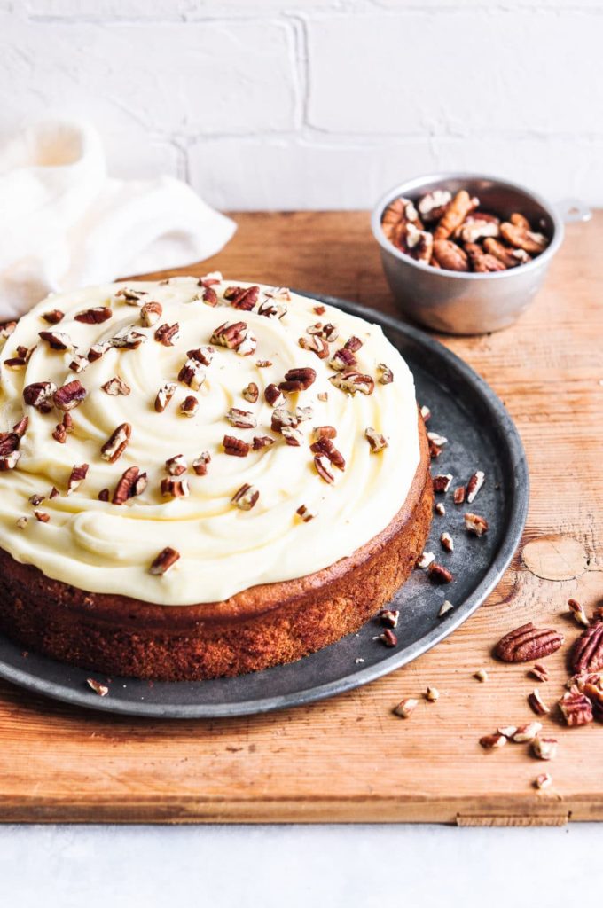 banana cake spread with cream cheese frosting and chopped pecans on metal tray on wooden board