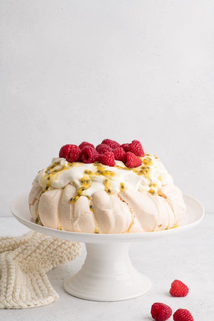 pavlova on white cake stand with whipped cream, passionfruit and raspberries