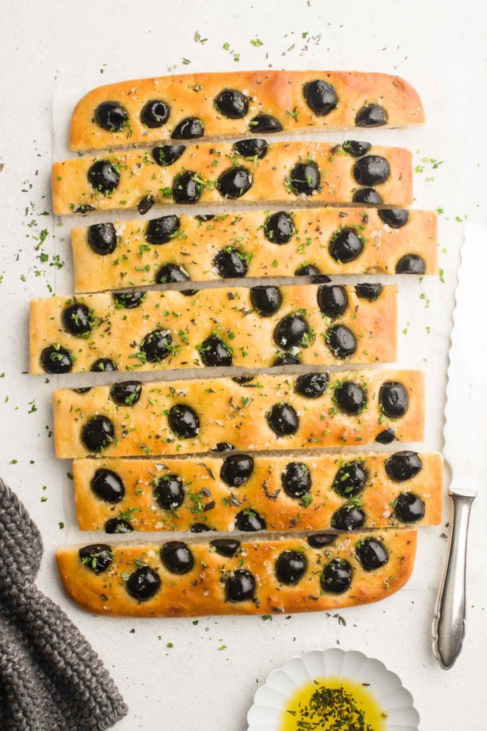 focaccia with herbs and olives with bread knife and grey cloth
